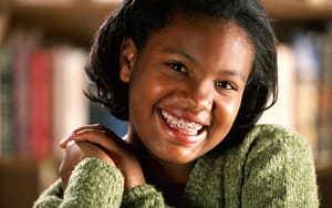 A young girl has fun with braces while smiling in front of a bookshelf.